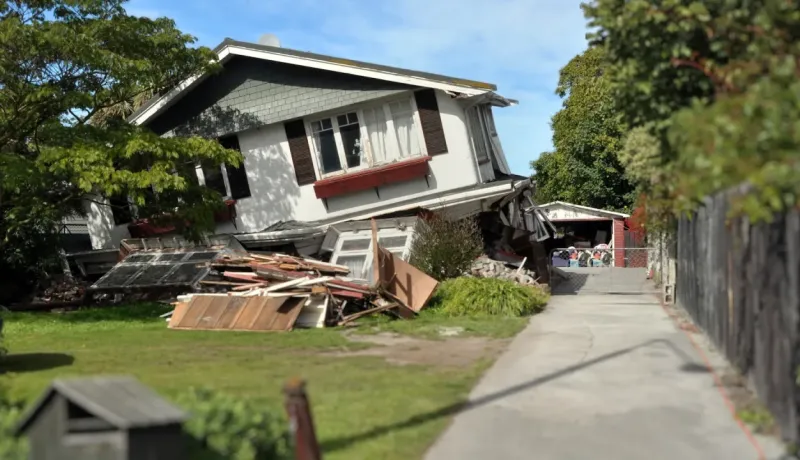 Maison inclinée et endommagée, probablement après un séisme, avec des débris empilés dans la cour avant.