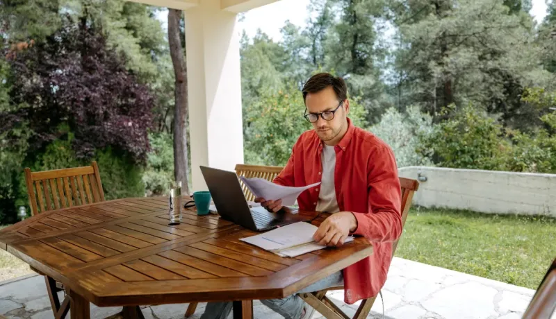 Homme en chemise rouge travaillant sur ordinateur portable et documents à une table en bois, en extérieur sous un auvent.