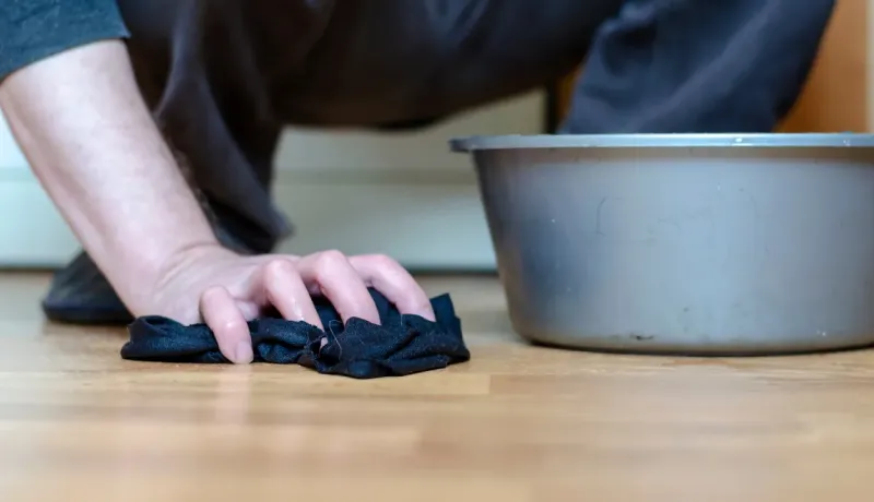 Gros plan sur une main nettoyant le sol en bois avec un chiffon noir, près d'une bassine d'eau.