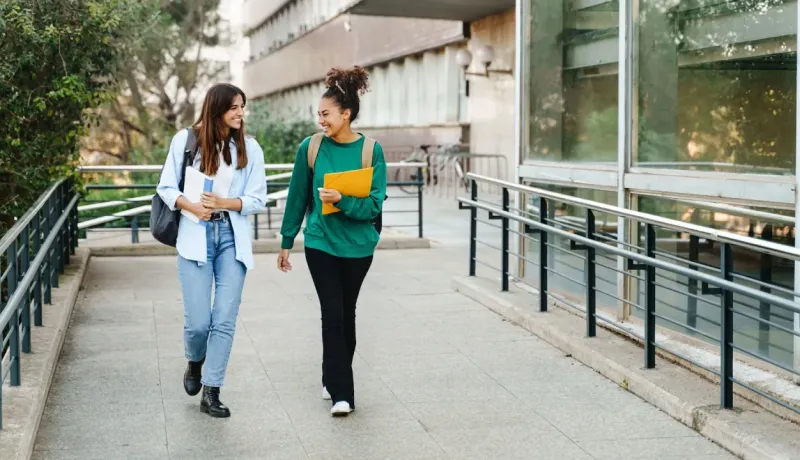 Deux étudiantes souriantes et décontractées marchent et discutent sur une passerelle du campus après les cours.