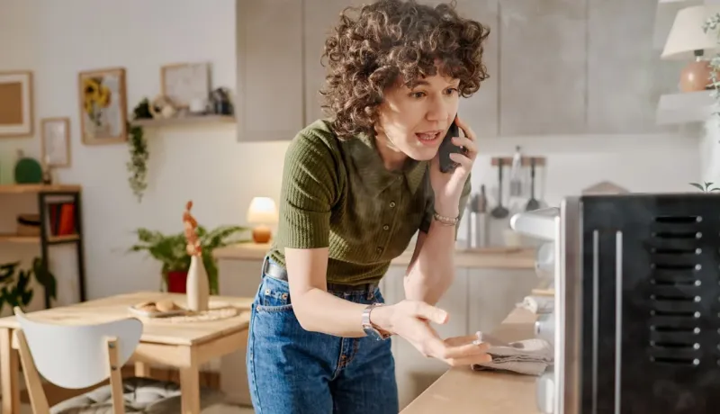 Femme agitée au téléphone dans sa cuisine, pointant du doigt de la vapeur s'échappant d'un appareil défectueux.