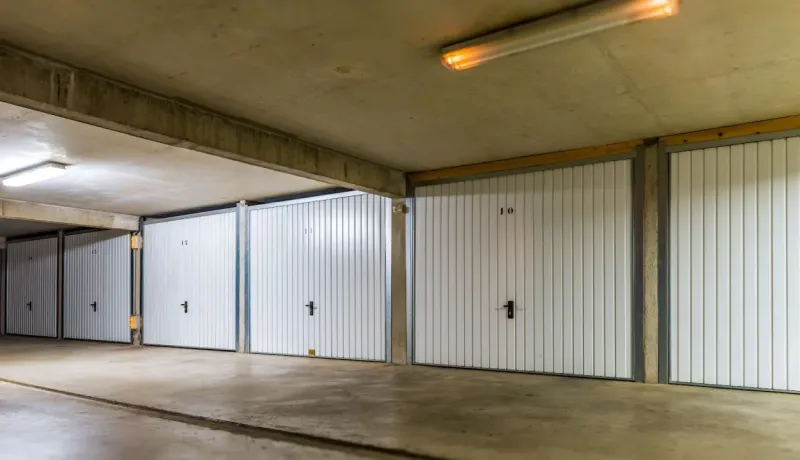 Rangée de boxes de garage individuels, numérotés et fermés par des portes blanches, dans un parking souterrain.