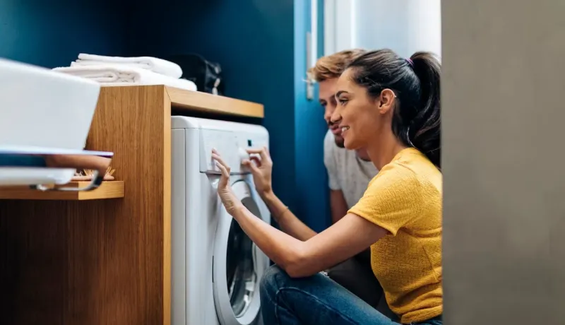 Couple souriant accroupi, réglant ensemble le programme d'un lave-linge dans une buanderie moderne.