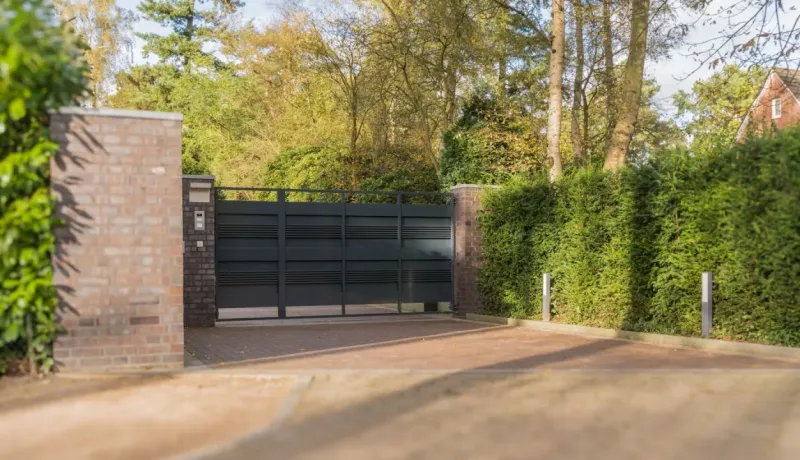 Grand portail métallique motorisé, flanqué de piliers en briques, donnant sur une allée et des haies.