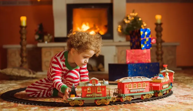 Petite fille en pyjama rayé jouant avec un train électrique de Noël devant une cheminée allumée.