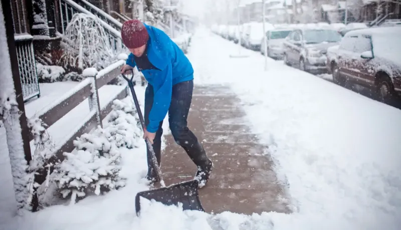 Personne déneigeant un trottoir envahi par la neige, dans une rue urbaine avec des voitures garées.