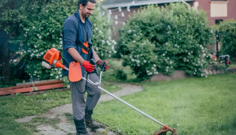 Homme utilisant une débroussailleuse pour couper l'herbe dans le jardin, portant des gants et un harnais de sécurité.