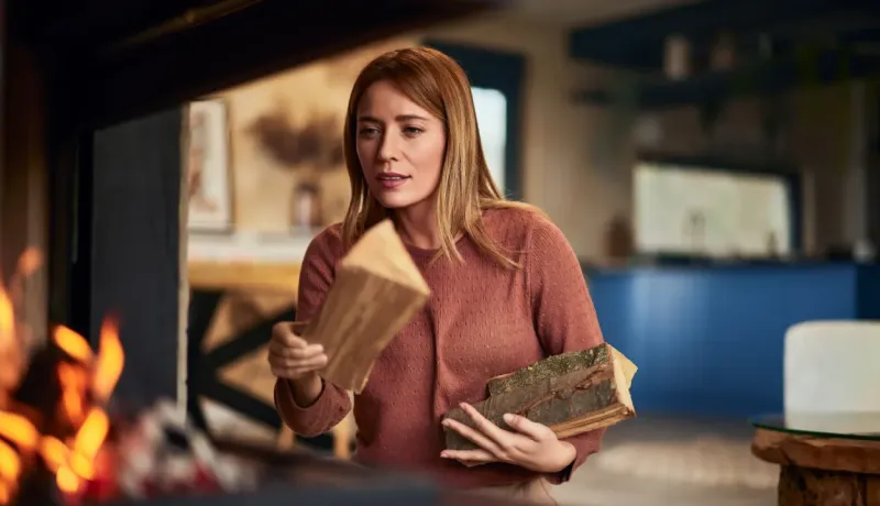 Femme aux cheveux blonds alimentant une cheminée ou un poêle avec des bûches dans un intérieur moderne et chaleureux.