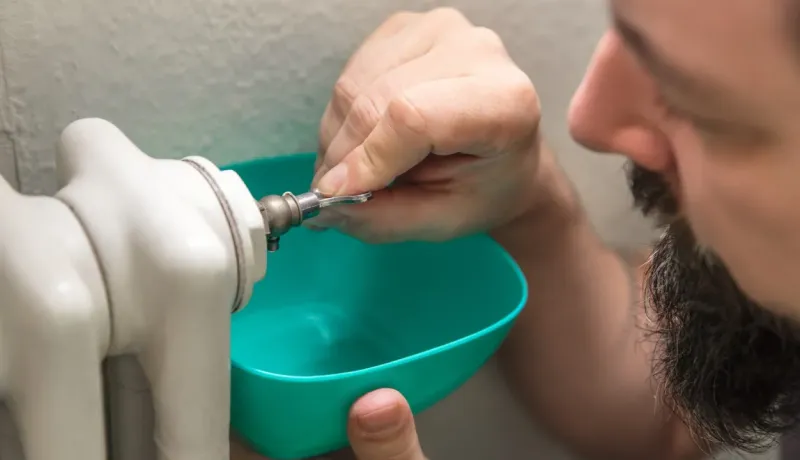 Gros plan sur un homme purgeant un radiateur avec une clé, tenant un bol turquoise pour recueillir l'eau.