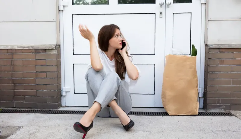 Une femme assise sur le pas de sa porte parle au téléphone, avec un sac d'épicerie en papier à côté d'elle.