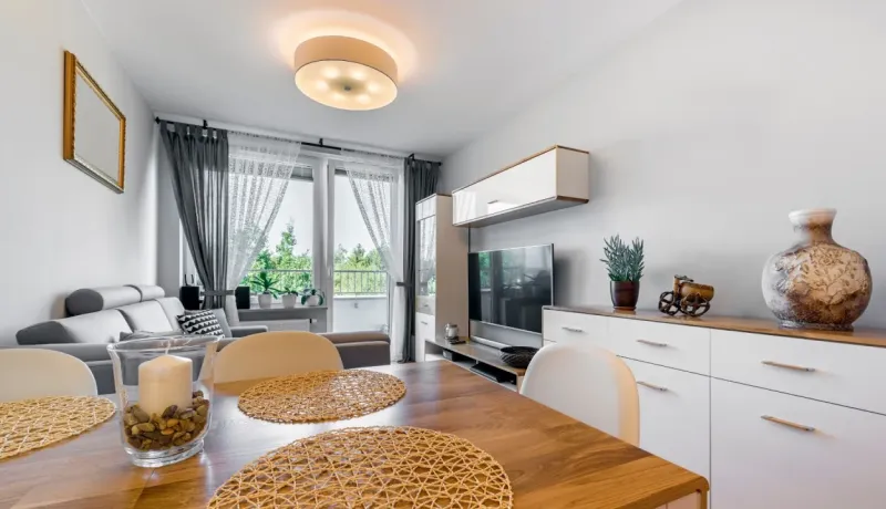 Vue intérieure d'un salon-salle à manger moderne aux murs blancs avec une table en bois clair, des chaises et un balcon vitré.