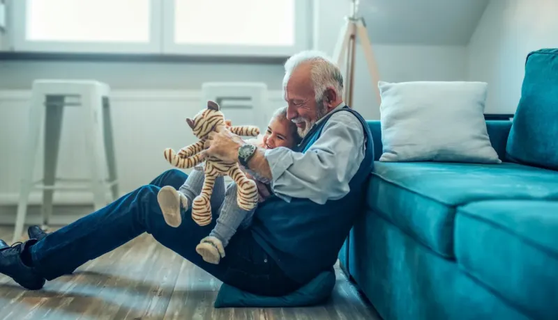 Un grand-père barbu et une petite fille rient ensemble sur le sol, jouant avec un chat en peluche tigré, près d'un canapé turquoise.