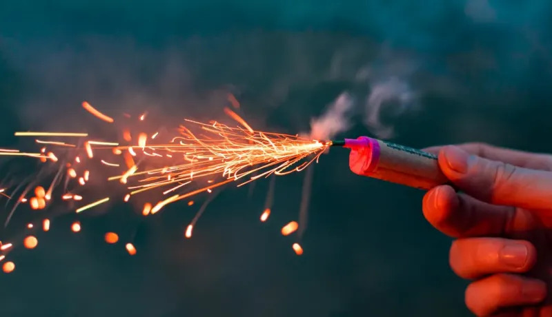 Gros plan sur une main tenant un petit feu d'artifice allumé, projetant des étincelles lumineuses dans l'obscurité.