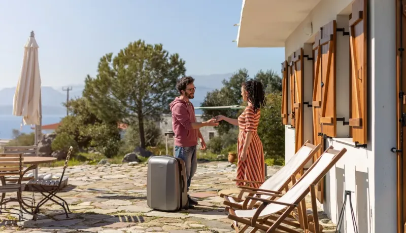 Un couple en vacances fait l'enregistrement. L'homme, avec une valise, reçoit une clé de la femme devant une villa.
