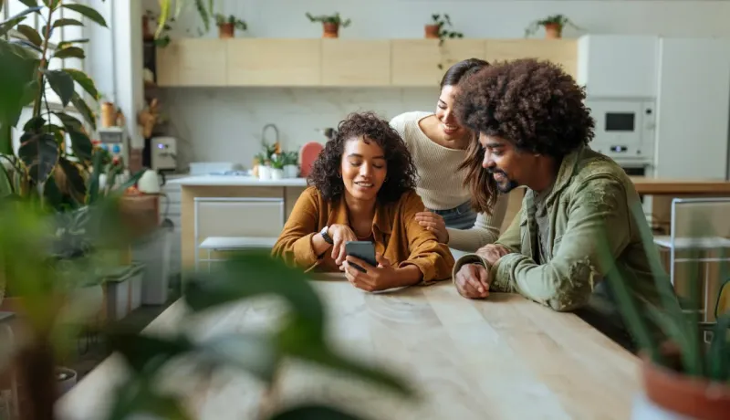 Trois jeunes amis sourient en regardant ensemble quelque chose sur un smartphone à table dans une cuisine lumineuse.