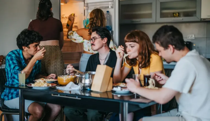 Un groupe d'amis d'âge universitaire mange des nouilles autour d'une table dans une cuisine, tandis que deux femmes sont au frigo.