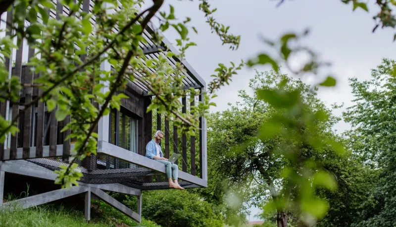 Un homme barbu travaille sur un ordinateur portable, assis sur le rebord d'un balcon en bois d'une maison moderne au milieu des arbres.