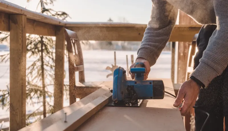 Gros plan sur les mains d'un homme qui coupe une planche de bois avec une scie circulaire sur une terrasse ensoleillée et enneigée.