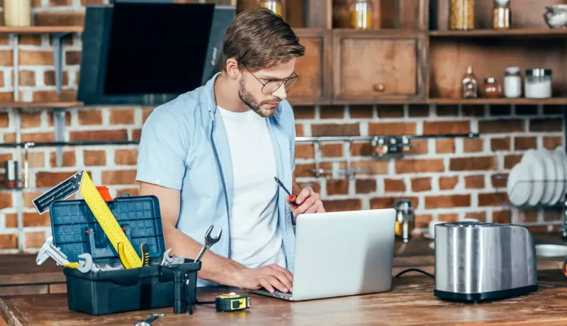 Un homme avec des lunettes utilise un ordinateur portable sur un plan de travail, à côté d'une boîte à outils ouverte, pour une réparation.