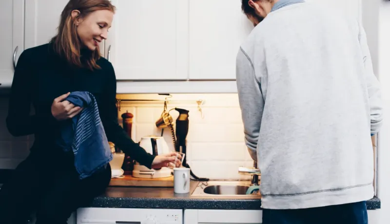 Un couple fait la vaisselle ensemble dans une cuisine : la femme s'appuie sur le comptoir en séchant une tasse, l'homme est devant l'évier.
