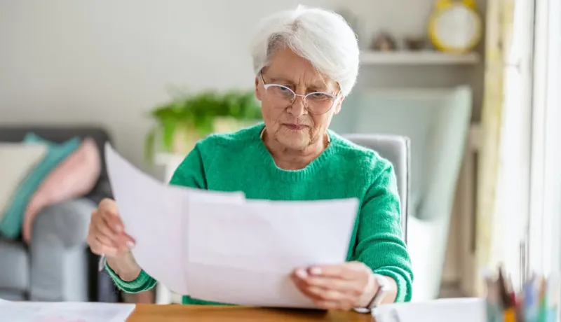Une femme âgée, concentrée, lit des documents ou des factures à sa table, portant un pull vert et des lunettes.