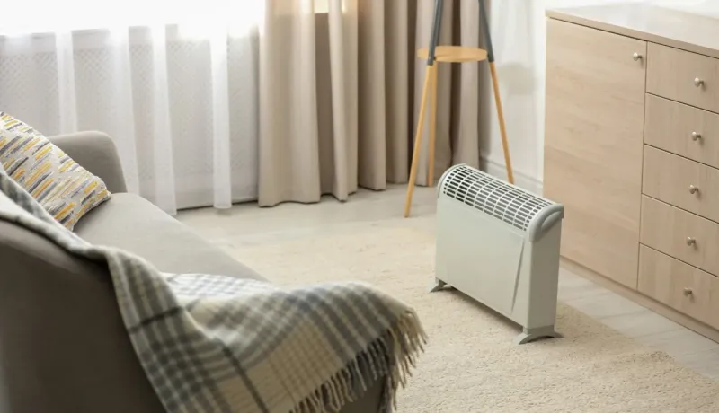Un petit radiateur électrique d'appoint chauffe un salon cosy avec un fauteuil, une moquette beige et des rideaux.