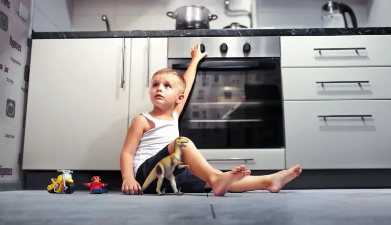 Un jeune garçon est assis sur le sol de la cuisine, jouant avec des jouets, et s'étire pour atteindre le bouton du four.