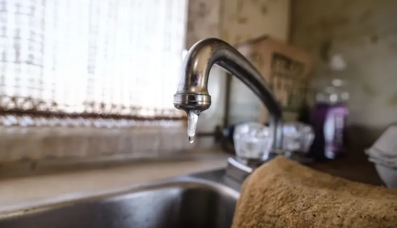 Gros plan sur un robinet de cuisine qui goutte, avec une seule goutte d'eau suspendue à l'extrémité, devant une fenêtre floue.