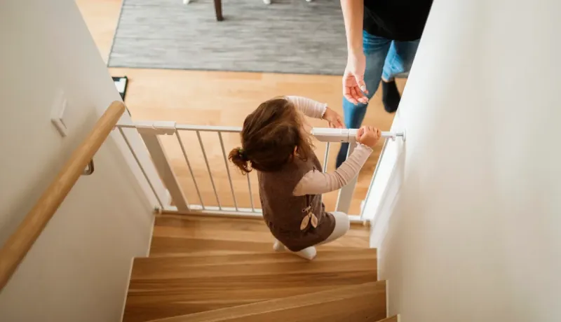 Vue de haut : une petite fille aux cheveux bruns est assise sur les marches en bois, agrippée à une barrière de sécurité pour escalier.
