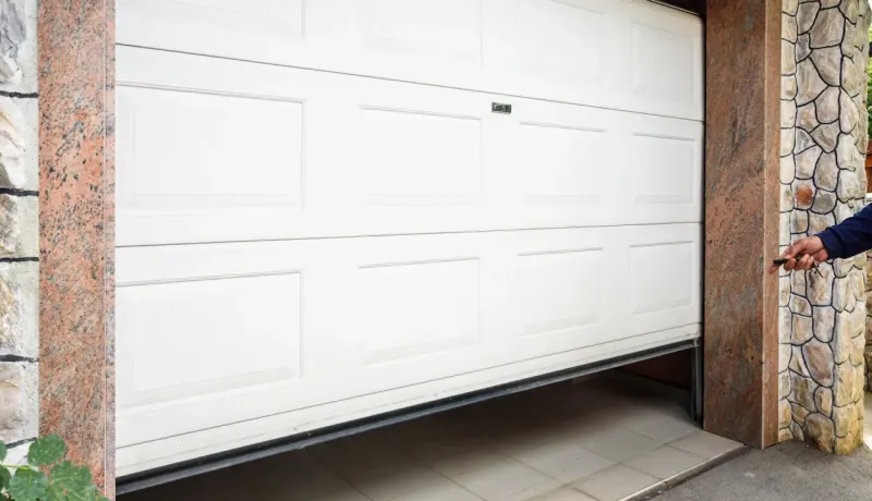 Une personne actionne un émetteur pour ouvrir un garage sectionnel blanc moderne, encadré par des pierres décoratives.