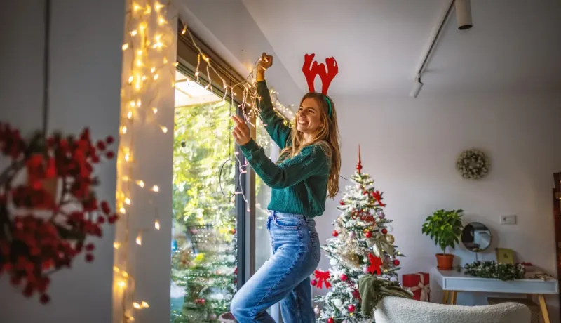 Une jeune femme joyeuse portant des bois de renne décore une fenêtre avec des guirlandes lumineuses à côté d'un arbre de Noël.