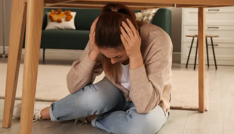Jeune femme s'abritant sous une table dans son salon, se protégeant la tête lors d'un exercice de tremblement de terre.