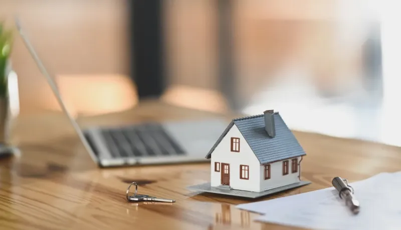 Gros plan sur une petite maquette de maison et des clés, posées sur une table de bureau près d'un ordinateur portable.