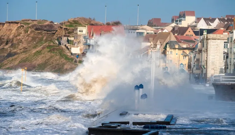 Une vague puissante s'écrase sur une digue, inondant la promenade devant des maisons côtières.