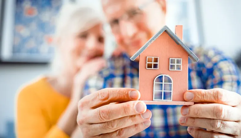 Gros plan sur un couple de personnes âgées souriant tenant ensemble une petite maquette de maison rose.