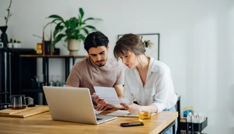 Un couple se penche sur un ordinateur portable, l'homme pointe un document : ils gèrent leurs finances ou lisent un contrat.