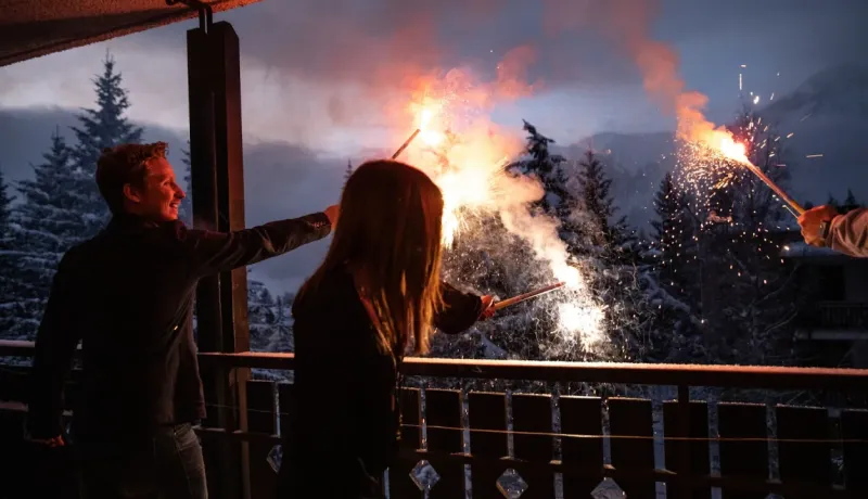 Un couple sur un balcon enneigé en montagne tient des cierges magiques, célébrant la nuit contre un paysage d'arbres.