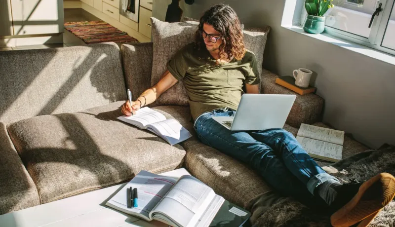 Un homme aux cheveux longs et aux lunettes est assis sur un canapé, étudiant ou travaillant avec un ordinateur portable et des livres.