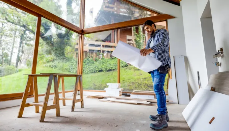 Un homme barbu en chemise à carreaux examine un plan dans une pièce en construction avec de grandes baies vitrées.