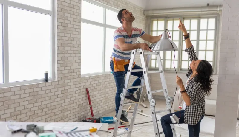 Un couple installe un luminaire. L'homme sur une échelle et la femme le guide, dans une pièce aux murs de briques.