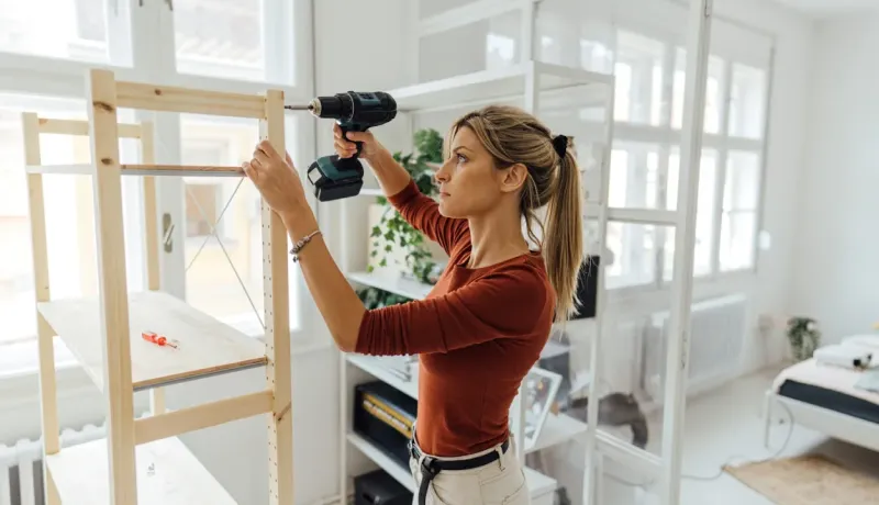 Une femme assemble une étagère en bois à l'aide d'une perceuse sans fil dans un salon lumineux.