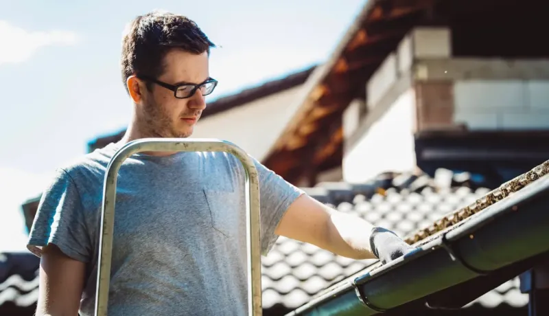 Un homme en lunettes et T-shirt gris est sur une échelle et nettoie la gouttière extérieure d'une maison.