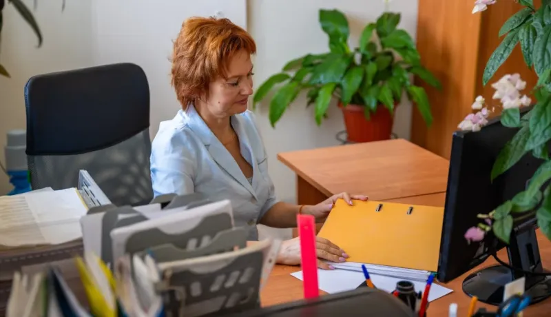 Une femme rousse est assise à un bureau, consultant un dossier jaune, entourée de plantes et de papeterie.