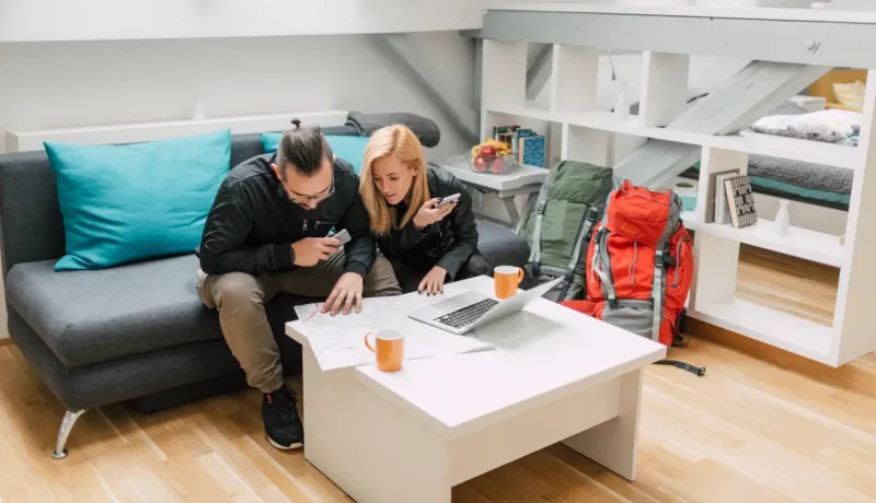 Un couple planifie un voyage, consultant une carte sur une table basse avec un ordinateur portable et des sacs à dos.