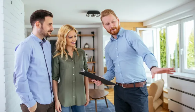 Un agent immobilier en chemise bleue montre l'intérieur d'un appartement à un jeune couple souriant.