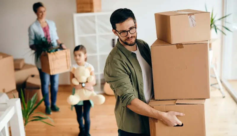 Un déménagement. Un homme transporte des cartons lourds, tandis qu'une femme et une enfant portent d'autres objets.
