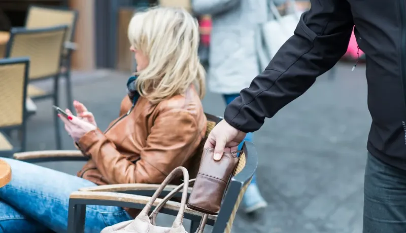 Un voleur dérobe un portefeuille brun posé sur une chaise, derrière une femme assise dans un café.