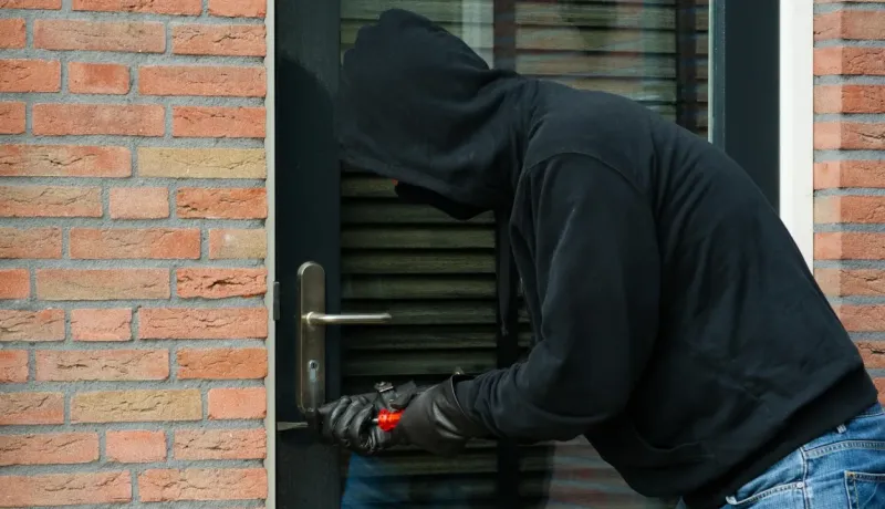 Un cambrioleur masqué, vêtu de noir, tente de forcer une serrure de porte avec un outil.