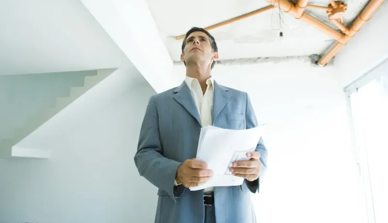 Agent immobilier en costume gris examine une propriété en construction avec des papiers à la main.
