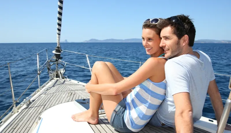 Un couple souriant assis sur le pont d'un voilier naviguant en pleine mer sous un ciel bleu.